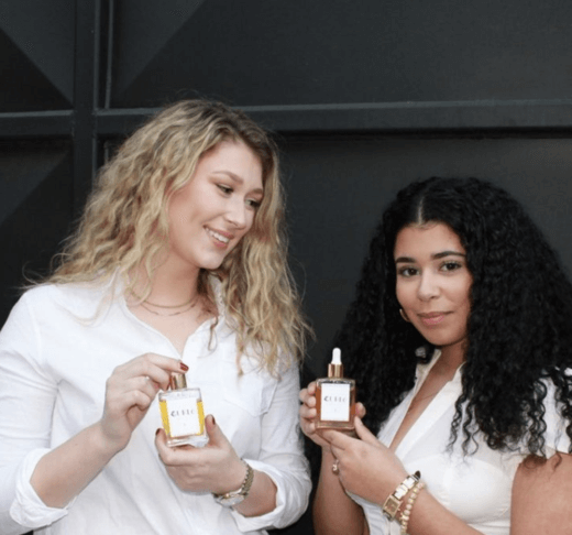 blonde curly haired girl smiling holding a curly hair oil to the left of a brunette curly haired Egyptian girl holding another curly hair oil both wearing white shirts 